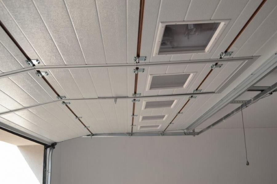 Biała segmentowa brama garażowa z oknami, widok od wewnątrz, widoczne prowadnice i elementy mocujące, brama częściowo otwarta, oświetlenie garażu.