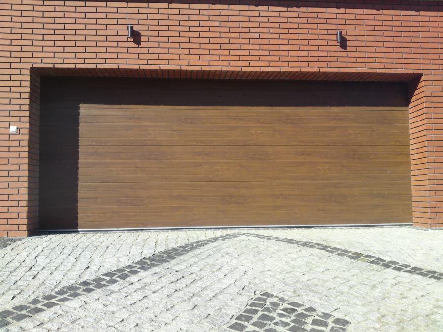 Zamknięta, brązowa brama garażowa segmentowa z imitacją drewna, wkomponowana w ceglany budynek. Podjazd wyłożony kostką brukową.