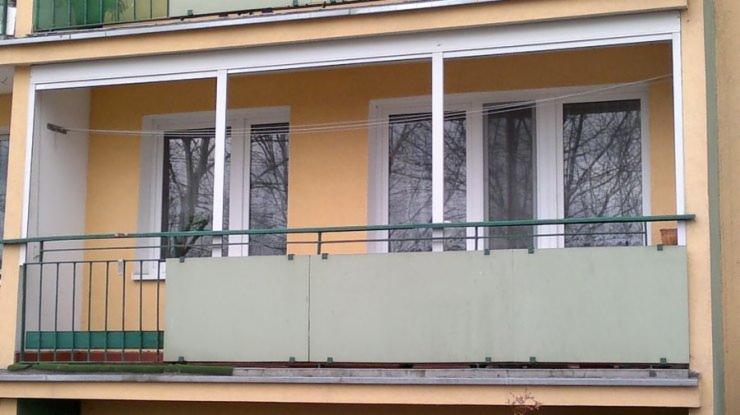 Zabudowany balkon z białymi ramami okiennymi i zieloną balustradą, widok z zewnątrz, drzewa odbijające się w szybach.
