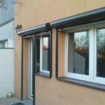 Otwarta okiennica zewnętrzna w brązowej ramie, na tle beżowej elewacji budynku. Widoczne okno z odbiciem nieba.