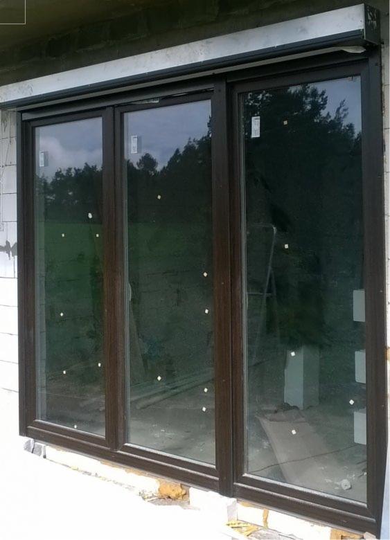 Zamknięte okno trzyskrzydłowe, brązowe ramy, widoczne zabezpieczenia na szybach. Zewnętrzna roleta zwinięta. Widok z zewnątrz, w tle zieleń.