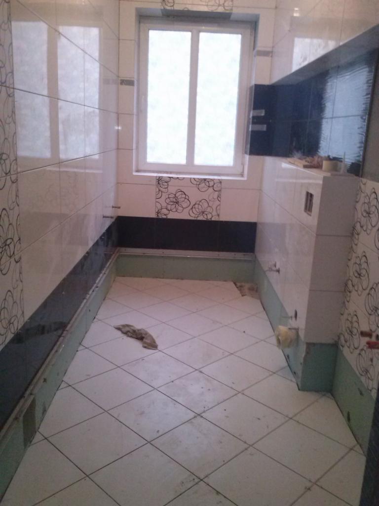 Wnętrze łazienki w trakcie remontu: widoczne płytki na ścianach z motywem kwiatowym, surowe podłoże podłogi z ułożonymi płytkami, okno z białymi ramami.