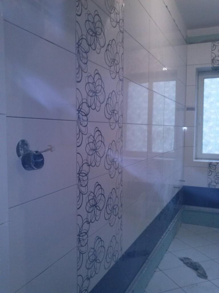 Wykończona ściana łazienki w Katowicach z białych płytek z motywem kwiatowym, widoczna instalacja wodna i fragment wanny.