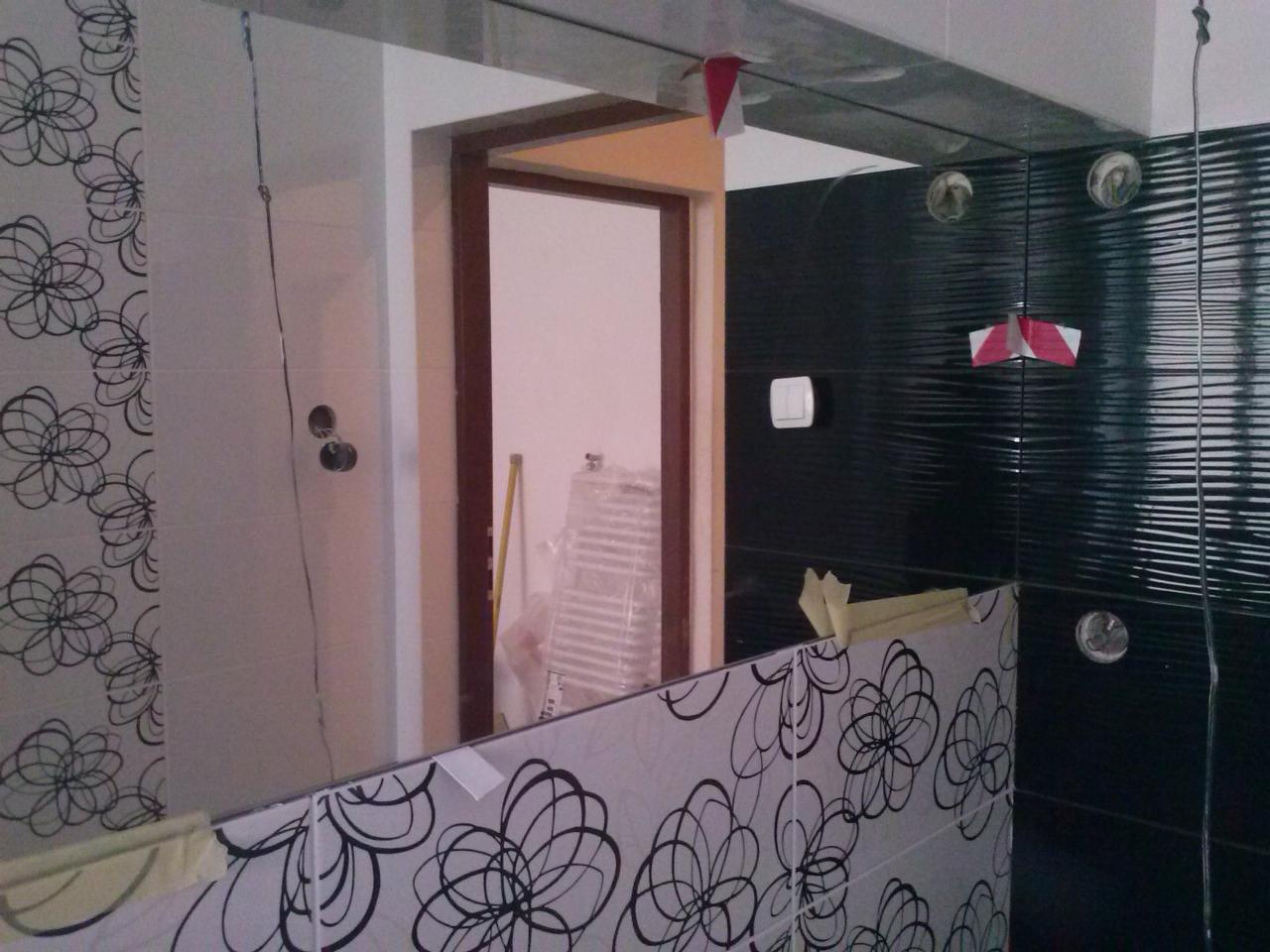 Częściowo wykończona łazienka z czarno-białymi płytkami, widoczne maskowanie i odbicie w lustrze. Etap prac wykończeniowych z akcesoriami w tle.