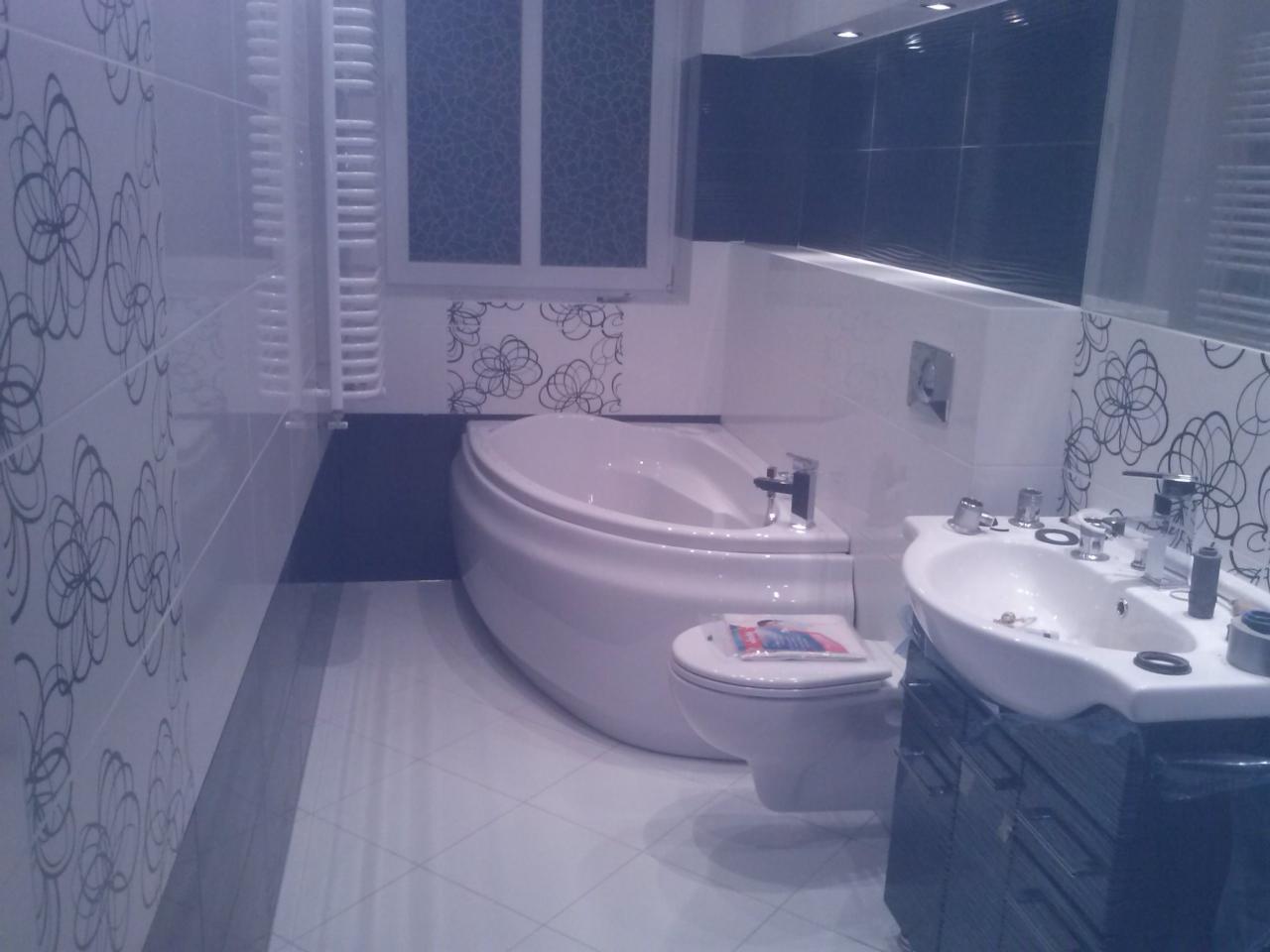 Nowoczesna łazienka z białą wanną, umywalką z szafką w kolorze grafitowym i płytkami w kwiatowy wzór. Widoczne okno z roletami i grzejnik.