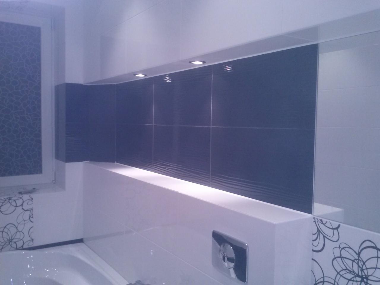 Wykończona łazienka z szarymi płytkami strukturalnymi, oświetleniem punktowym i dekoracyjnymi motywami kwiatowymi na białych płytkach. Lustro odbija fragment łazienki.