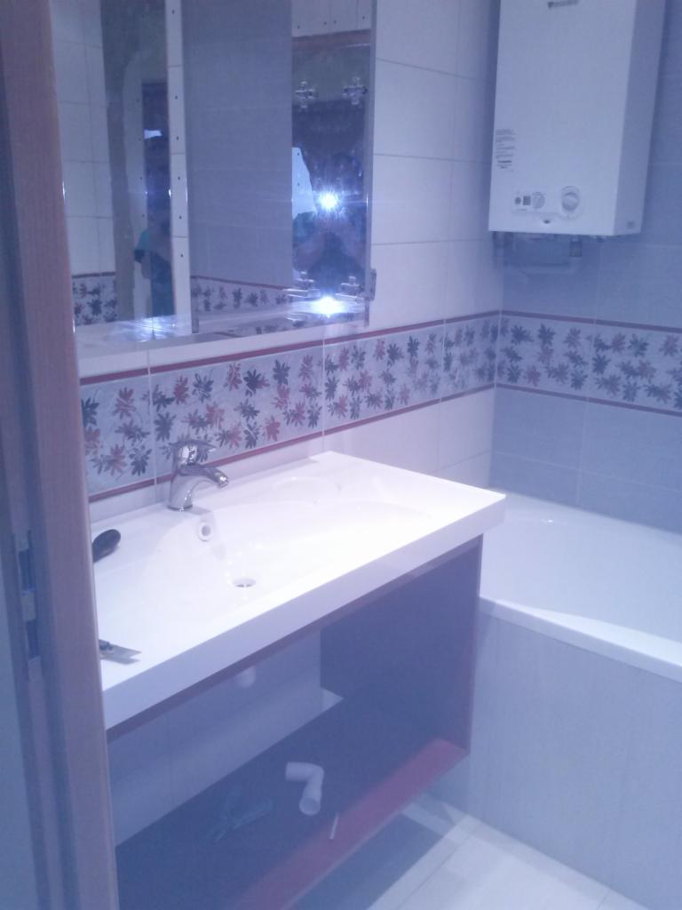 Wykończona łazienka z białą umywalką, baterią, lustrem i grzejnikiem. Płytki z subtelnym wzorem zdobią ściany. Ujęcie z perspektywy wejścia.