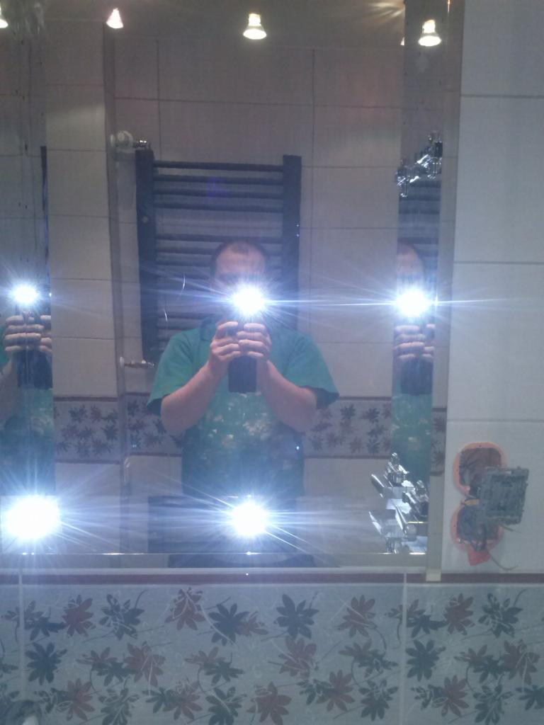 Odbicie osoby w lustrze łazienkowym z widoczną instalacją elektryczną i grzejnikiem drabinkowym. Widoczne płytki z motywem kwiatowym.