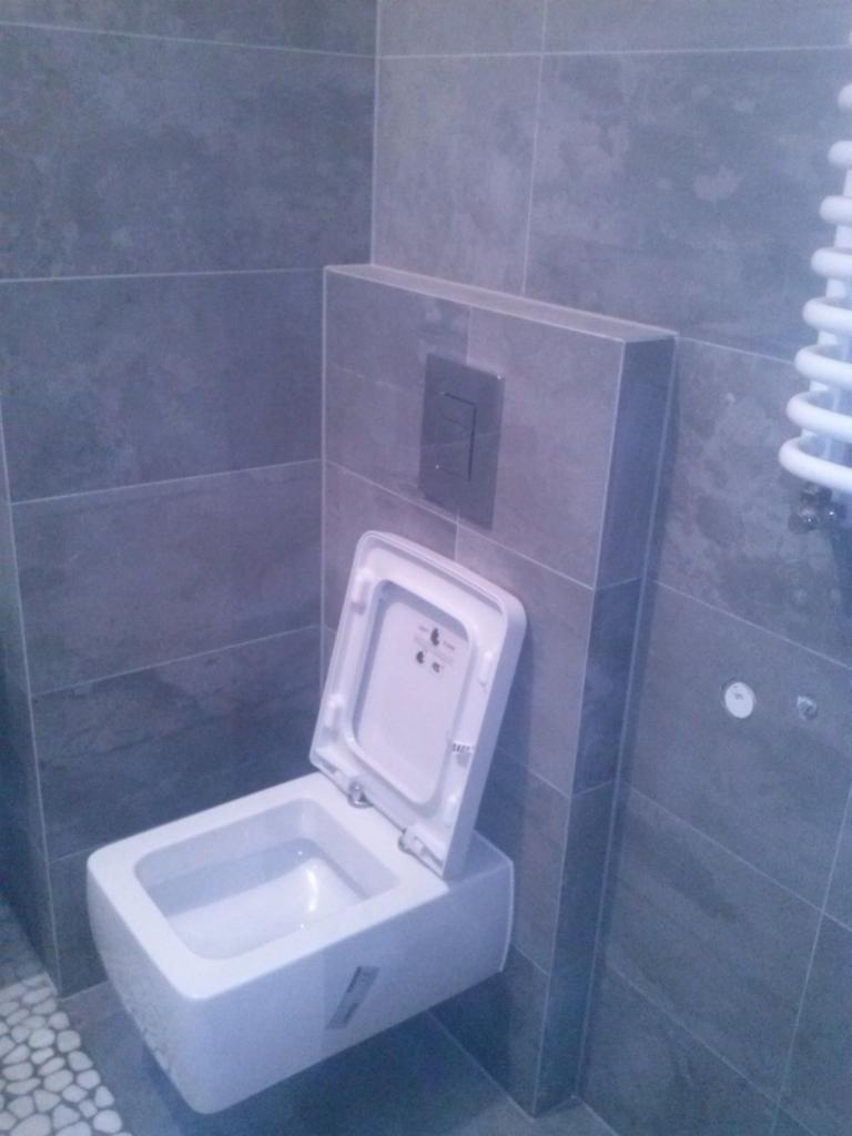 Nowoczesna, podwieszana toaleta z otwartą klapą w wykończonej łazience. Szare, duże płytki na ścianach i mozaika na podłodze. Widoczny grzejnik drabinkowy.