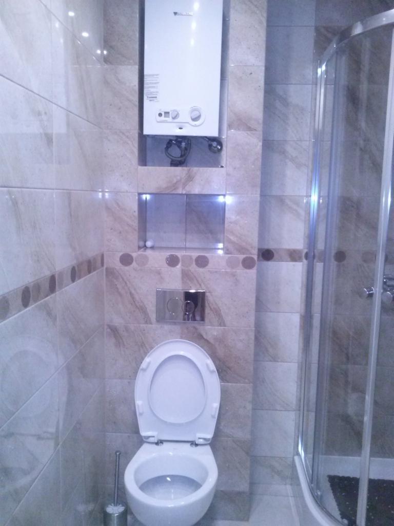 Wykończona łazienka z płytkami w odcieniach beżu i brązu. Widoczny bojler, toaleta i kabina prysznicowa z chromowanymi elementami. Nowoczesny design.