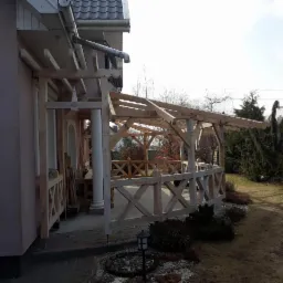 Konstrukcja drewnianej altany przylegającej do budynku, widoczne elementy ozdobne i krzyżowe wzmocnienia balustrady, na etapie montażu więźby dachowej.