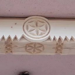 Detal zdobienia drewnianego elementu konstrukcyjnego z wyciętymi wzorami geometrycznymi: rozetami i ząbkami, widok z dołu na tle sufitu.