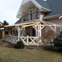 Konstrukcja drewnianej pergoli przy domu jednorodzinnym w trakcie budowy, widoczne rusztowania i narzędzia, z przodu ozdobne ogrodzenie w kształcie krzyży.