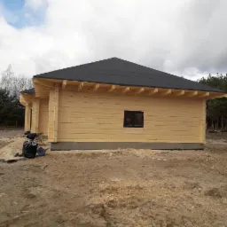 Surowy, drewniany dom z bali z czarnym dachem, widok z boku na tle lasu i pochmurnego nieba, z widocznymi elementami budowy i workami na śmieci.