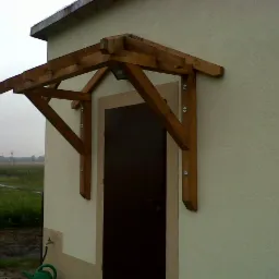 Drewniany zadaszenie nad drzwiami wejściowymi, przymocowane do ściany budynku, widok na pole w tle.