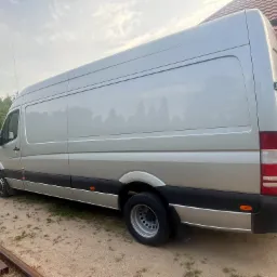 Srebrny bus dostawczy marki Mercedes na żwirowym podłożu, z widocznymi elementami zabudowy i fragmentem domu w tle. Długi rozstaw osi, wysoki dach.