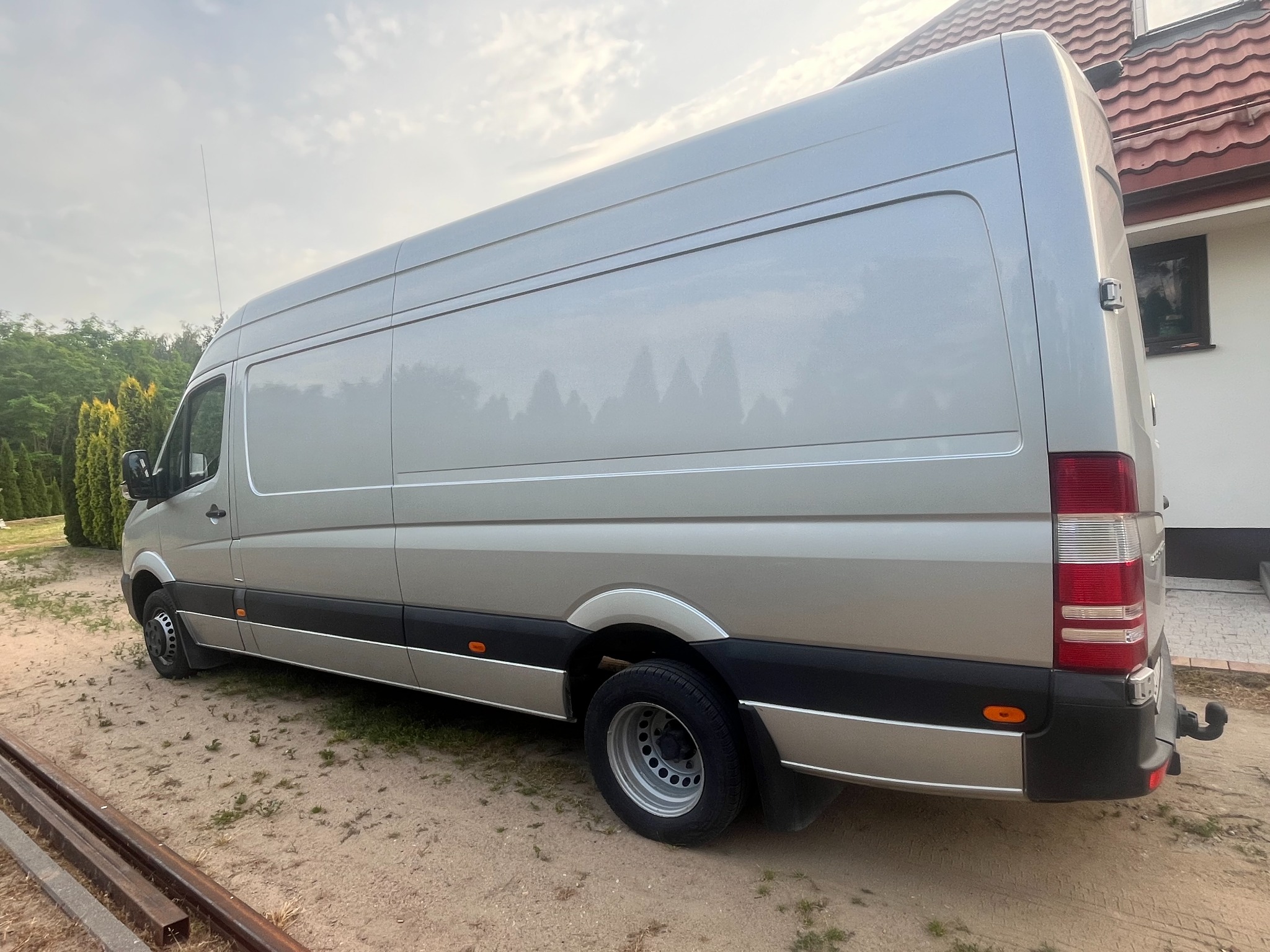 Srebrny bus dostawczy marki Mercedes na żwirowym podłożu, z widocznymi elementami zabudowy i fragmentem domu w tle. Długi rozstaw osi, wysoki dach.