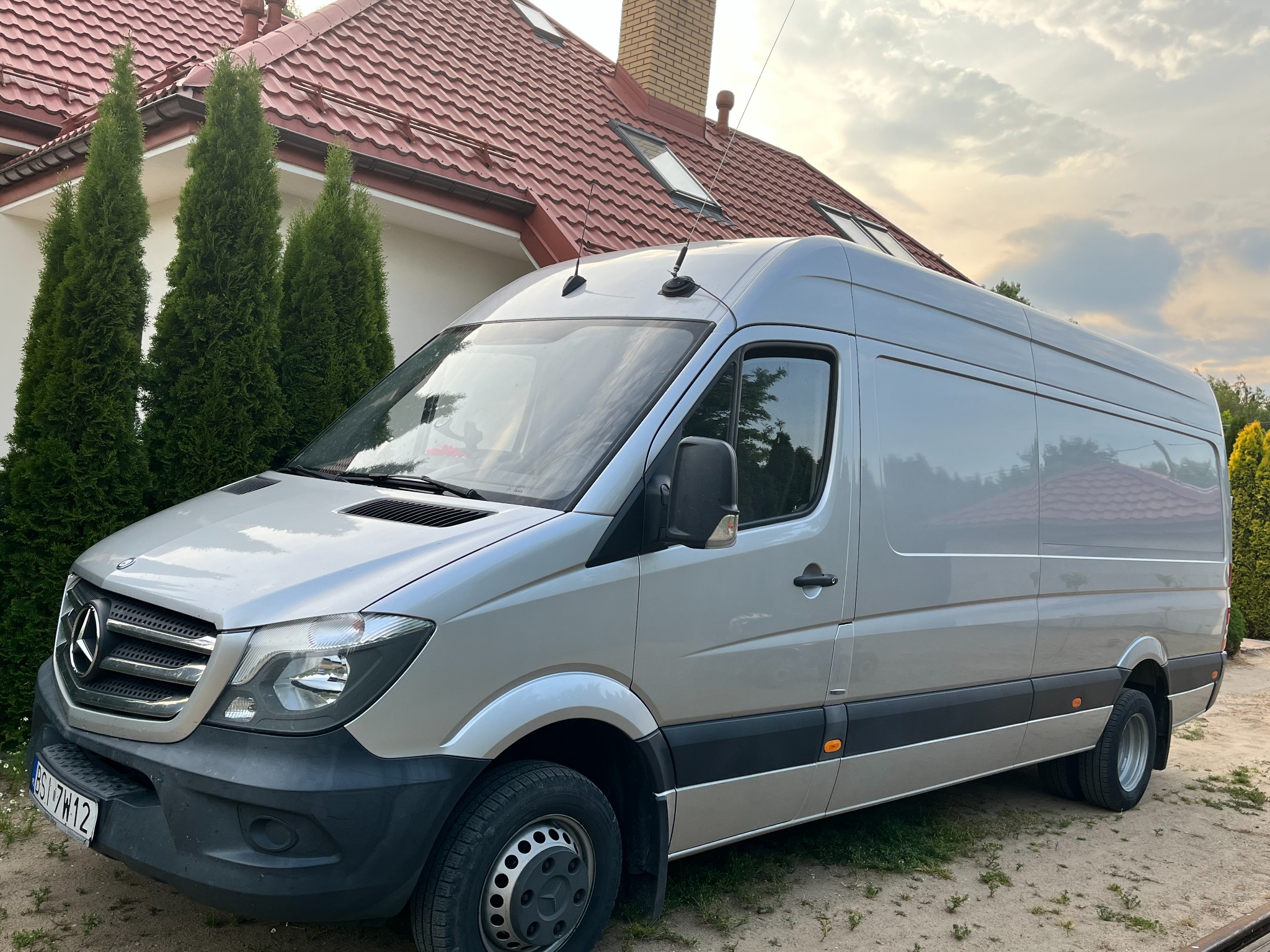 Srebrny bus Mercedes-Benz Sprinter, zaparkowany przed domem z czerwoną dachówką i zielonymi tujami. Widok z boku, lekko z przodu, na tle zachmurzonego nieba.