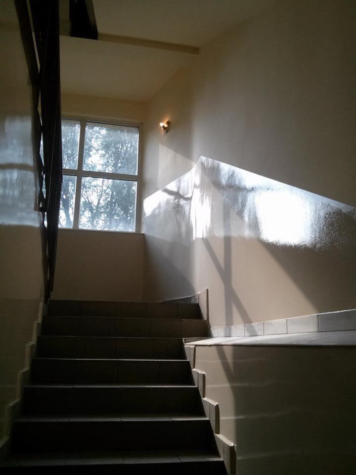 Klatka schodowa z drewnianą balustradą, oświetlona promieniami słońca wpadającymi przez okno. Widoczne schody z szarymi płytkami i jasne ściany.