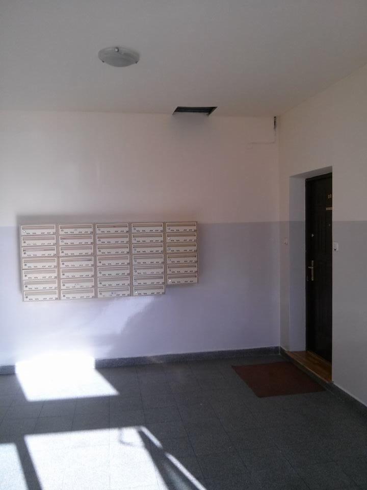 Wnętrze klatki schodowej z rzędem skrzynek pocztowych na ścianie, szara podłoga z płytek, widoczne drzwi wejściowe i oświetlenie sufitowe.