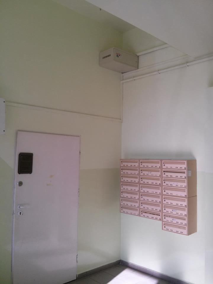 Klatka schodowa z metalową skrzynką na ścianie, obok drzwi i zestawu skrzynek pocztowych w bloku mieszkalnym. Widok na fragment korytarza.