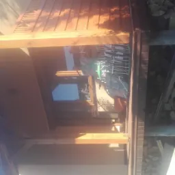 Drewniana konstrukcja zadaszenia tarasu w trakcie budowy, widoczne elementy więźby dachowej i przygotowane drewno na opał w tle.