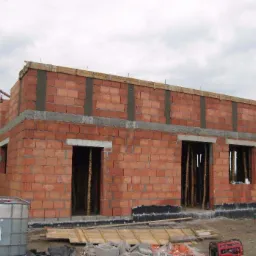 Budowa domu jednorodzinnego w stanie surowym otwartym, widoczne ściany z czerwonej cegły, otwory okienne i drzwiowe z drewnianymi stemplami, szalunek na stropie, teren budowy z elementami izolacji...