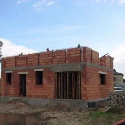 Wznoszenie murów parterowego domu z czerwonej cegły ceramicznej, widoczne zbrojenia nad otworami okiennymi, dwóch pracowników na dachu, palety cegieł obok budynku.