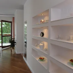 Białe, podświetlone wnęki w ścianie z ekspozycją dekoracyjnych przedmiotów, widok na salon z parkietem i balkonem. Nowoczesne wykończenie wnętrza.