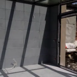 Nowoczesna, przeszklona weranda z szarymi, kwadratowymi płytkami na ścianach i podłodze, oświetlona promieniami słonecznymi wpadającymi przez szklany dach, widoczne kable elektryczne wystające ze...