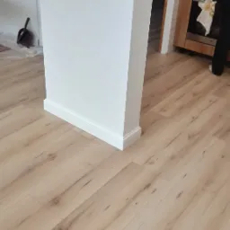 Jasne panele podłogowe ułożone wzdłuż białej ścianki działowej z listwami przypodłogowymi, widoczny fragment kuchni w tle