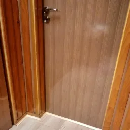 Zamknięte drzwi wewnętrzne z brązowego drewna z metalową klamką, przylegające do boazerii w odcieniach brązu i beżu, z widoczną białą listwą przypodłogową i fragmentem podłogi imitującej drewno.