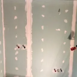 Ściana w trakcie prac przygotowawczych do wykończenia, widoczne płyty gipsowo-kartonowe z zaszpachlowanymi łączeniami, wystające rury instalacji wodnej w kolorach czerwonym i niebieskim,...