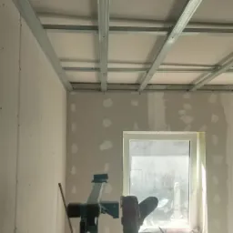 Stelaż sufitu podwieszanego z płyt kartonowo-gipsowych z widocznym podnośnikiem do płyt i narzędziami na parapecie okiennym w trakcie montażu.