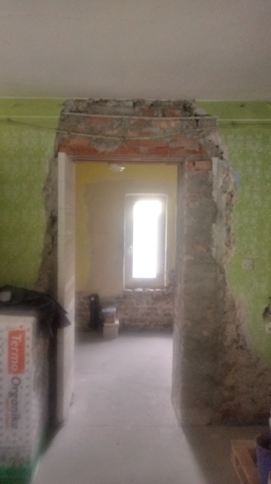 Demontaż nadproża drzwiowego w trakcie prac remontowych, odsłonięta cegła, widoczne ślady po starym tynku na zielonej ścianie.