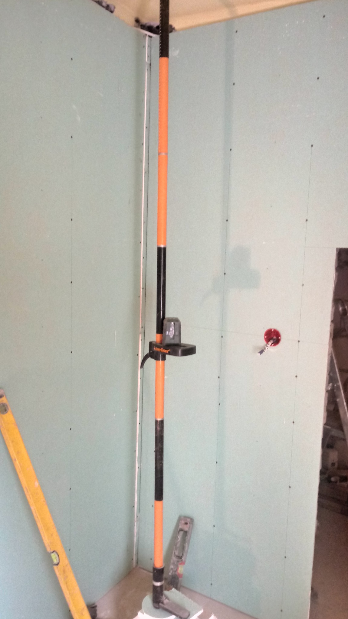 Montaż płyt gipsowo-kartonowych w narożniku pomieszczenia, widoczny laser krzyżowy na statywie, poziomica i otwór pod gniazdko elektryczne.