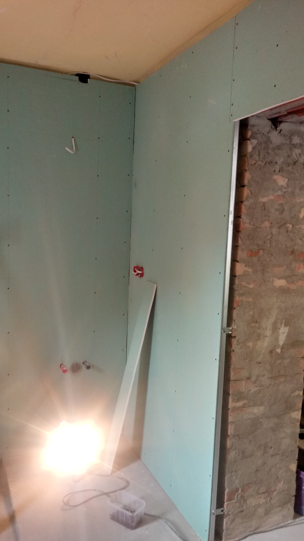 Wnętrze w trakcie montażu płyt gipsowo-kartonowych na ścianach, widoczne instalacje elektryczne, fragment ściany z cegły oraz lampa budowlana na podłodze.