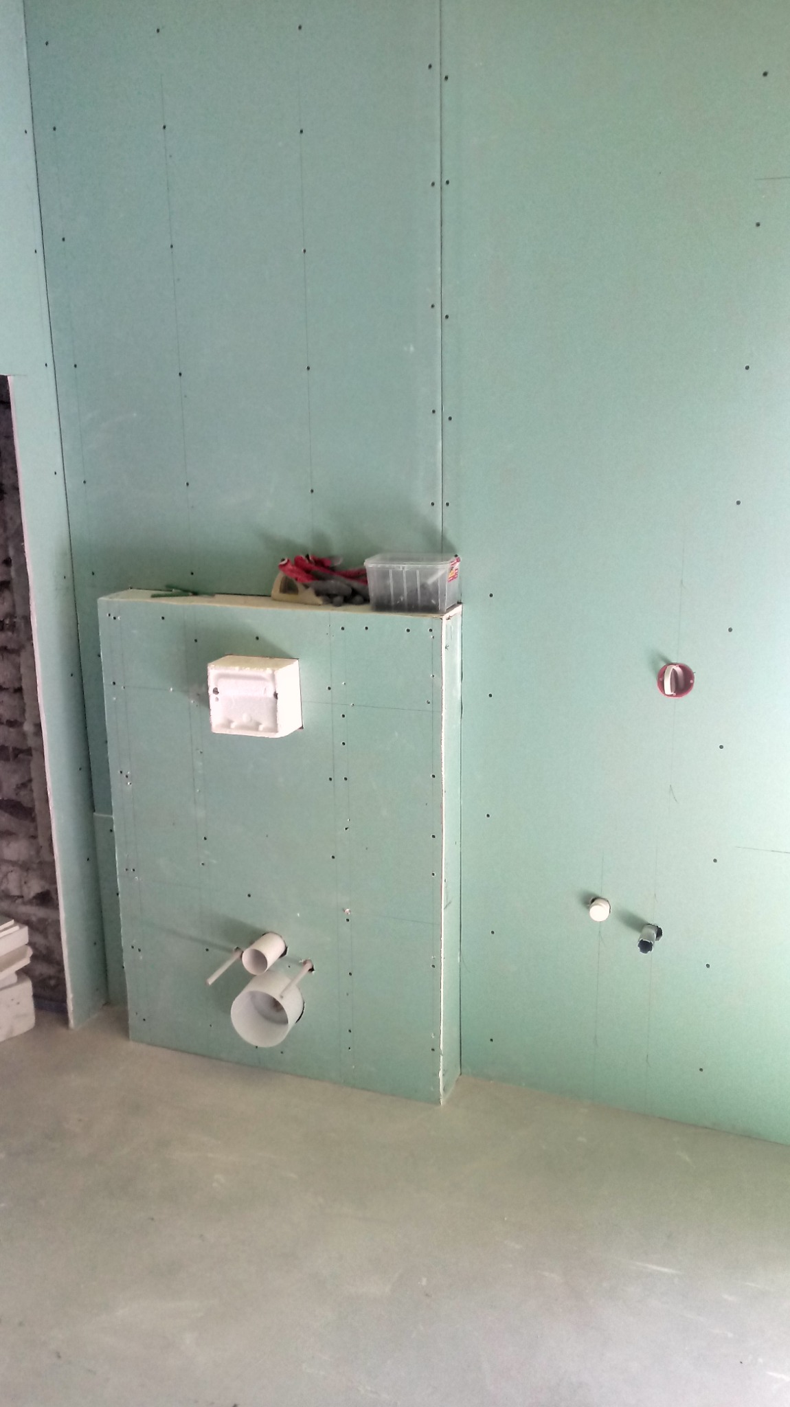 Ściany łazienki wykończone płytami gipsowo-kartonowymi, widoczne przygotowane instalacje wodno-kanalizacyjne, oraz narzędzia i pojemnik na górze zabudowy geberitu.