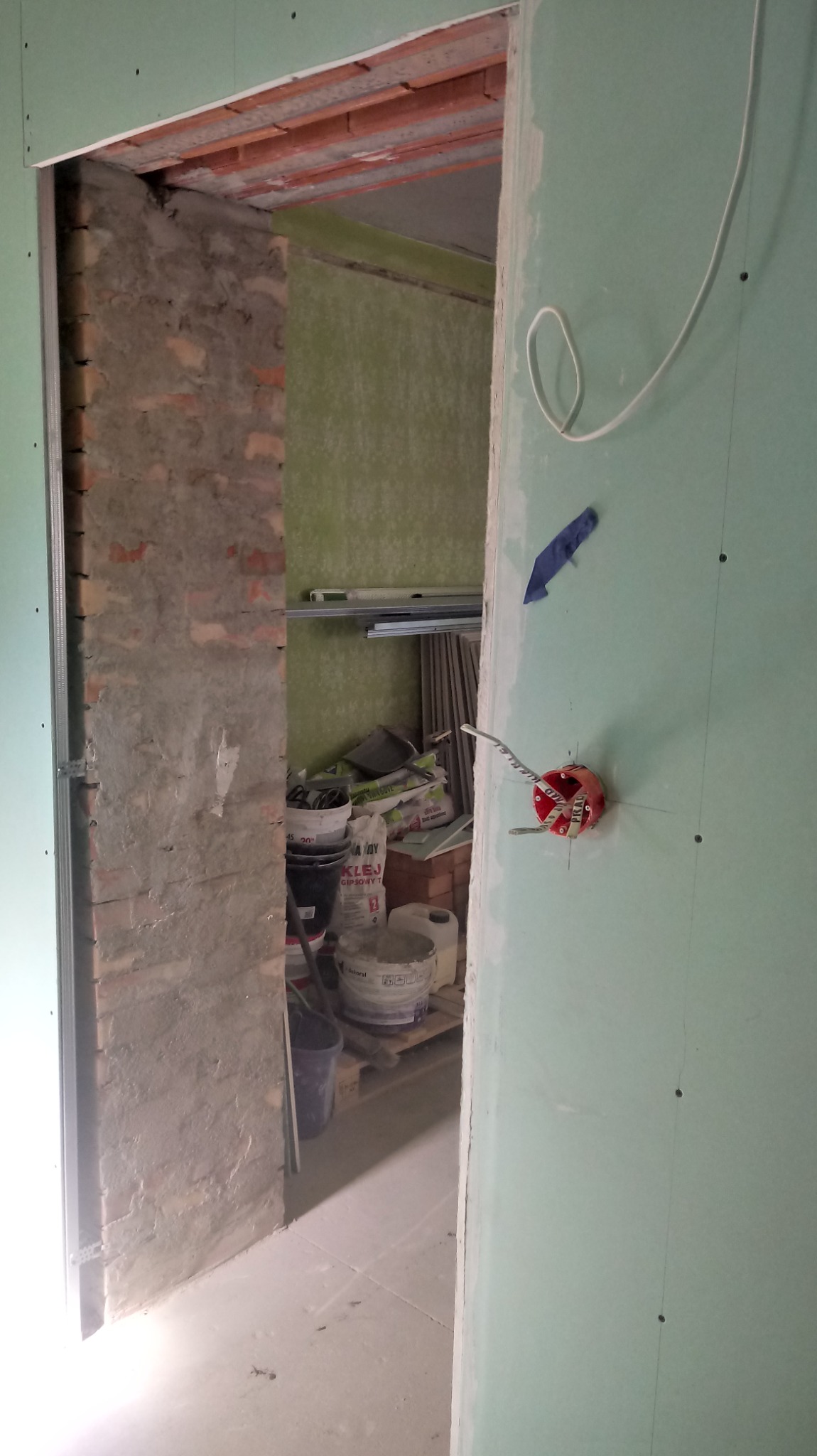 Ściana z płyty gipsowo-kartonowej w trakcie montażu, widoczna puszka elektryczna z wystającymi przewodami, fragment ściany z cegły i materiały budowlane w tle.