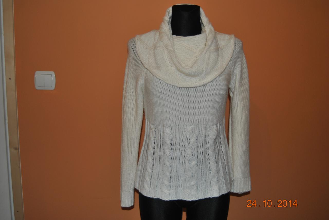 Kremowy sweter z grubym, ozdobnym splotem i szerokim golfem, zaprezentowany na manekinie na tle pomarańczowej ściany. Widoczny włącznik światła i data.