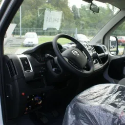 Wnętrze nowego Fiata Ducato z folią ochronną na siedzeniu, widoczny kokpit, kierownica i otwarte drzwi, w tle inne pojazdy na parkingu.