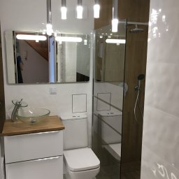 Zdunek Remonty Dariusz Zdunek - Nowoczesna łazienka z szklaną kabiną prysznicową, umywalką na blacie z drewnianym wykończeniem i podwieszanymi lampami. Widoczne białe płytki z delikatnym wzorem.