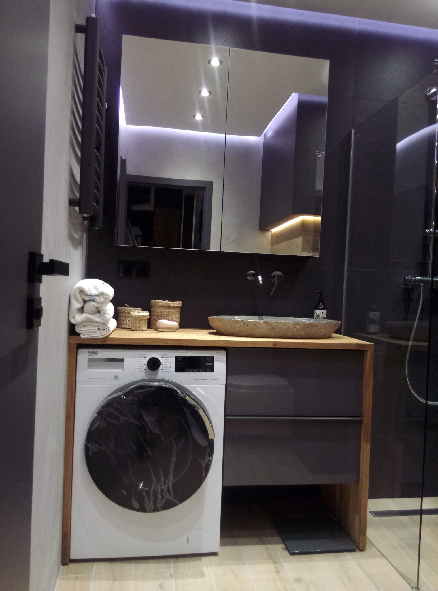 Nowoczesna łazienka w Koszalinie z pralką pod blatem umywalki, szare płytki, oświetlenie LED i lustro z szafką. Minimalistyczny design.