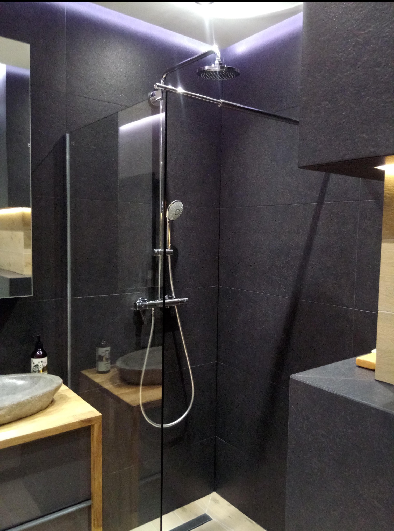 Nowoczesna kabina prysznicowa z deszczownicą i szklaną ścianką, wykończona ciemnymi płytkami imitującymi kamień, z podświetleniem LED i kamienną umywalką.