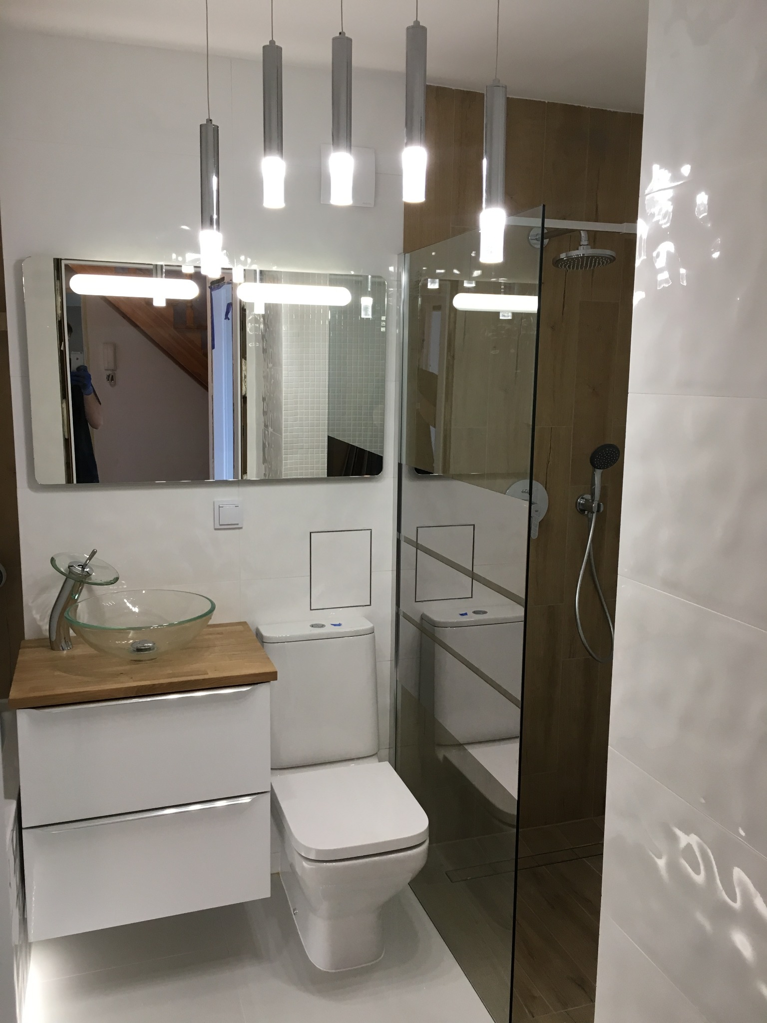 Nowoczesna łazienka z szklaną kabiną prysznicową, umywalką na blacie z drewnianym wykończeniem i podwieszanymi lampami. Widoczne białe płytki z delikatnym wzorem.