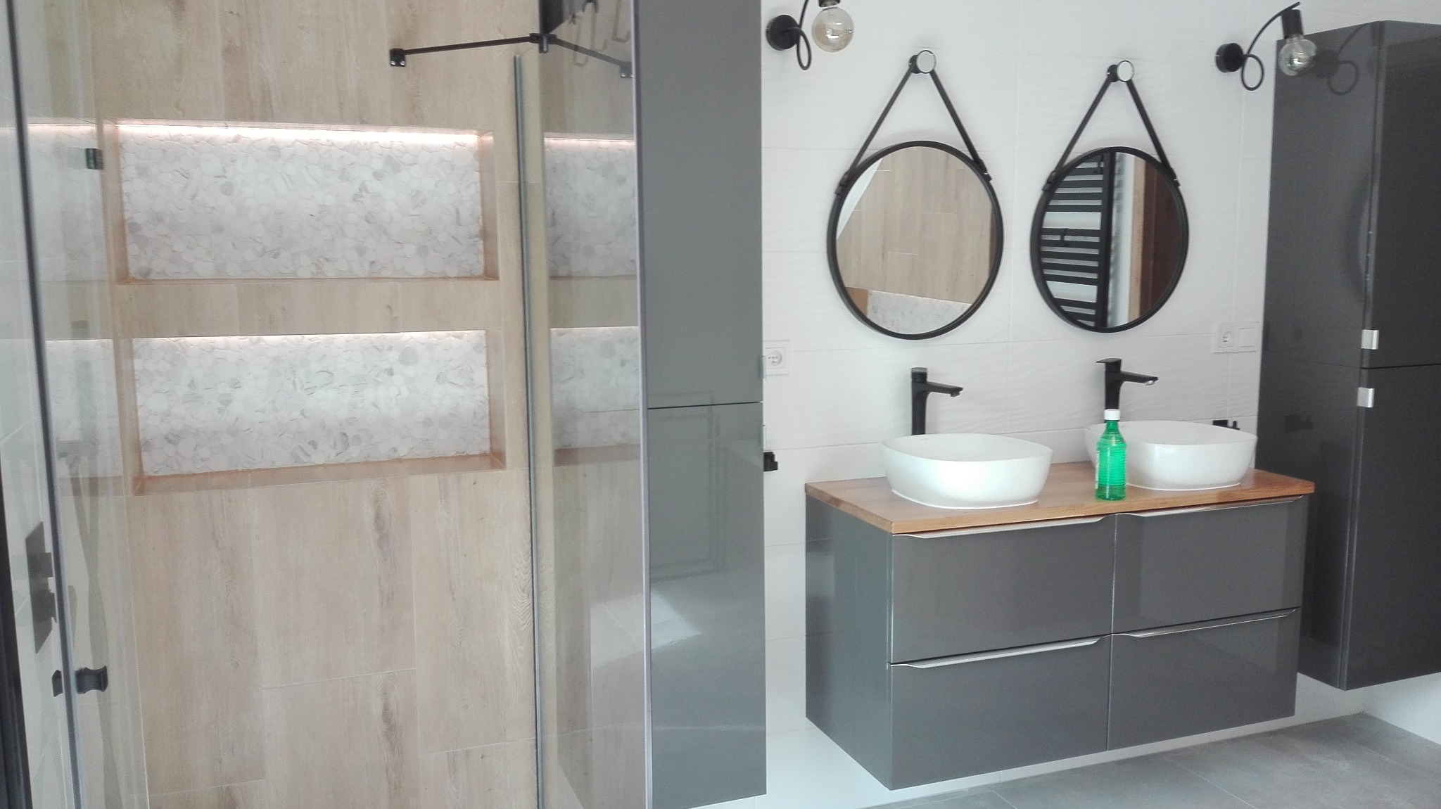 Nowoczesna łazienka z dwoma okrągłymi lustrami, umywalkami nablatowymi i kabiną prysznicową z podświetlanymi wnękami wykończonymi mozaiką.