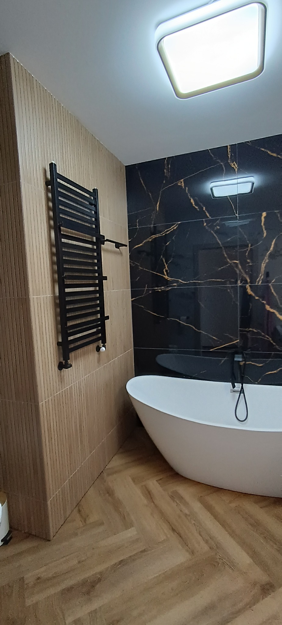 Nowoczesna łazienka z wolnostojącą wanną, czarnymi płytkami imitującymi marmur ze złotymi żyłami i grzejnikiem drabinkowym na ścianie z drewnopodobnych paneli.