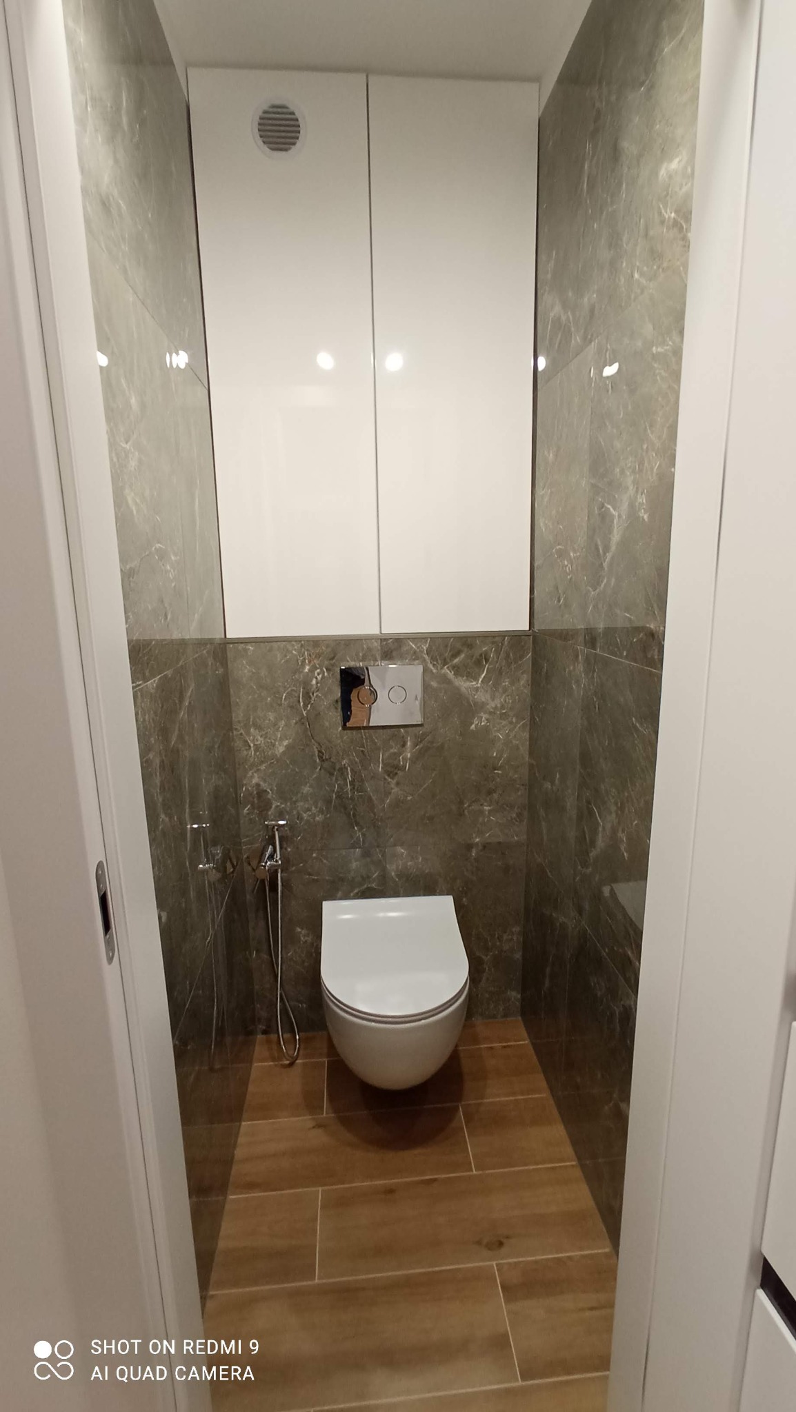 Nowoczesna toaleta w łazience z marmurowymi płytkami, podwieszaną miską WC i drewnopodobną podłogą. Białe szafki wiszące z wentylacją.