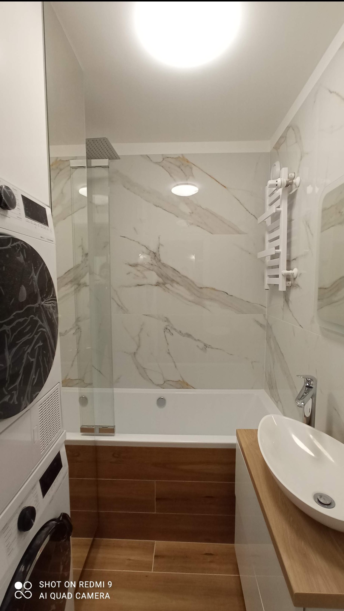 Nowoczesna łazienka z wanną obudowaną drewnopodobnymi płytkami, marmurowe płytki na ścianach, szklany parawan nawannowy i biała umywalka nablatowa.