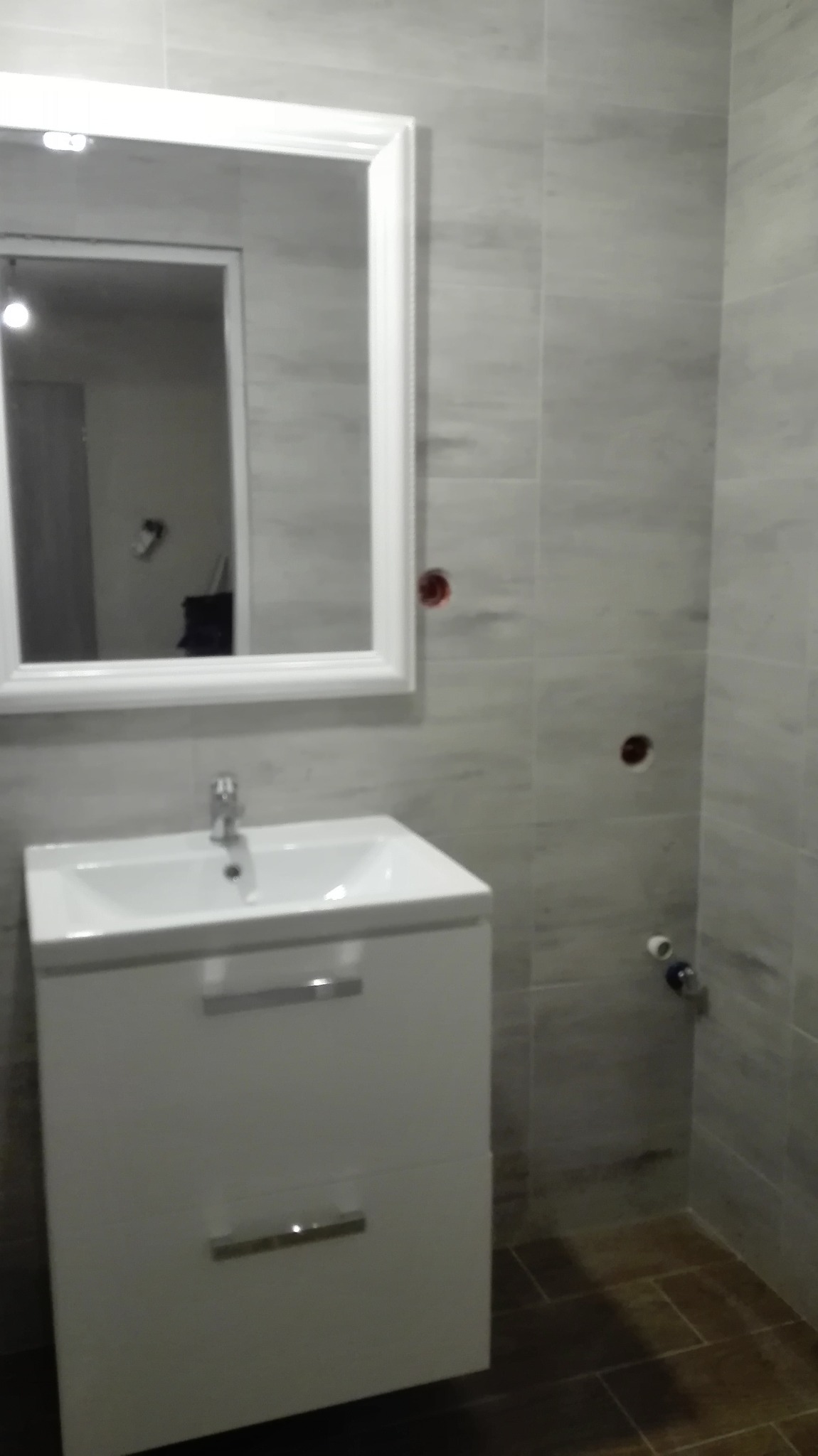 Umywalka z szafką i lustrem w białej ramie na tle szarych płytek. Widoczne niedokończone punkty instalacji elektrycznej i wodnej. Brązowe płytki podłogowe.